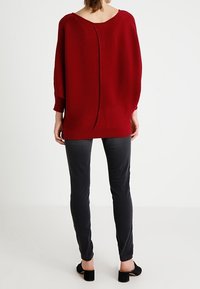 Punainen neulepusero, jossa on leveä veneen kaula-aukko ja takasaumadetail, yhdistettynä tummanharmaisiin slim fit -housuihin ja mustiin matalakorkoisiin kenkiin.