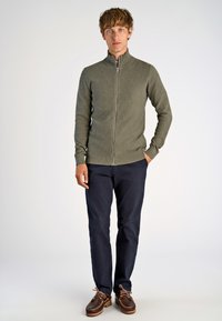 Maglione zip-up color khaki con una superficie texture, abbinato a pantaloni blu navy e scarpe da barca marroni. L'insieme presenta un colletto alla coreana e un design aderente.
