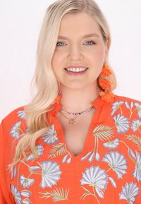 Orange Bluse mit floralem Stickerei in Blau und Grün. Auffällige Accessoires sind eine mehrfarbige Perlenkette und Ohrringe in Form von orangefarbenen Blumen.