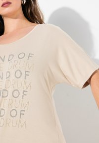 Lys beige kortærmet top lavet af blødt stof, med et gentaget tekstdesign i metallisk sølv og guld accenter.