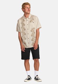 Camisa de manga corta, color beige, con bordado floral, bolsillo en el lado izquierdo del pecho, pantalones cortos oscuros, calcetines blancos y zapatillas negras.