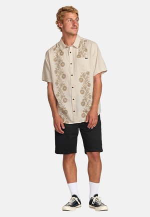 Camicetta beige a maniche corte con ricami floreali, taschino sul lato sinistro del petto, pantaloni corti scuri, calzini bianchi e sneakers nere.
