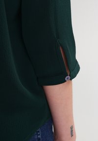 Dunkelgrüner, strukturierter Blusärmel mit kleinem, mit Knöpfen versehenem Schlitz, getragen von einer Person, die ein Unterarm-Tattoo und einen Teil von Jeans zeigt.