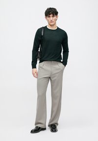 Dark green textured sweater, light cream collar, gray patterned trousers, black shoes, holding a small black bag, standing against a white background.