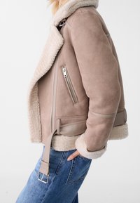 Tansuede schapenvachtjack met een grote kraag, zijritsen en een tailleband. Zachte textuur met contrasterende crèmekleurige voering.