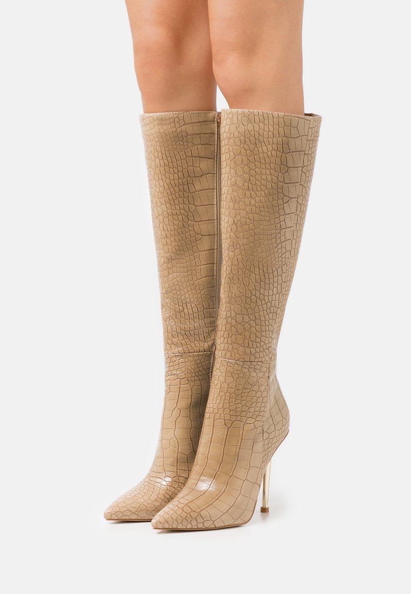 Bottes hautes beiges à talons aiguilles avec un motif effet crocodile, dotées d'un bout pointu et d'un talon stiletto métallique fin. Texture lisse.