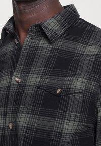 Black and dark grey plaid button-up shirt with chest pocket, worn by a person with dark skin.