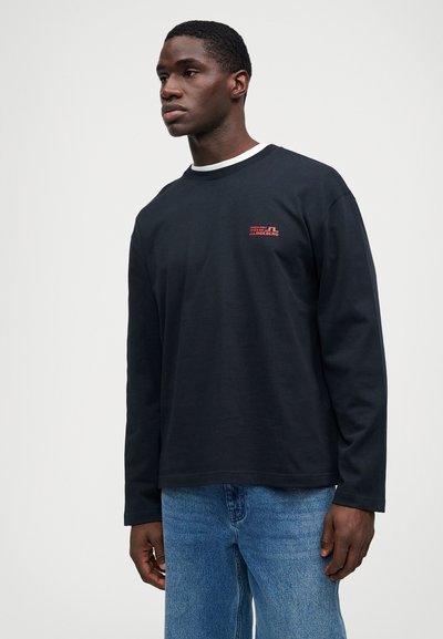 Egy férfi fekete hosszúujjú pólót visel piros logóval a mellkasán, és kék farmernadrágot, egy sima fehér háttér előtt áll.