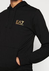 Černá mikina s kapucí vyrobená z měkkého materiálu s přední kapsou. Disponuje zlatým logem EA7 na levém prsou a kapucí se šňůrkou.