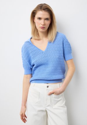 Joven mujer con suéter de manga corta de textura azul claro y pantalones blancos de cintura alta, de pie contra un fondo blanco.