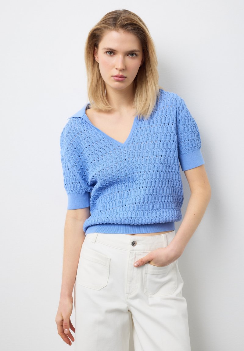 Jonge vrouw in lichtblauwe, gestructureerde trui met korte mouwen en witte, hooggetailleerde broek staat tegen een witte achtergrond.