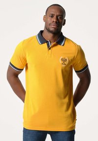 Geel polo-shirt met contrasterende marineblauwe kraag en manchetten, voorzien van een geborduurd logo en tennismotief op de borst. Zachte katoenmateriaal.