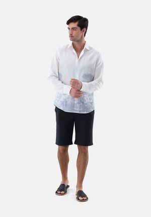 Mann trägt ein weißes langärmeliges Hemd mit hellblauem Muster, schwarze Shorts und schwarze Sandalen und steht mit verschränkten Händen.