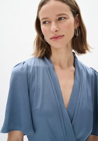 Hellblaues Wickelkleid mit kurzen, ausgestellten Ärmeln. Strukturierter Stoff mit einem subtilen Muster. Tiefes V-Ausschnitt-Design, minimalistischer Stil.