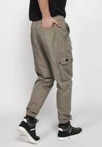 Khaki cargo hlače z elastičnimi manšetami, več žepi in vrvico v pasu, v kombinaciji s črnimi visokimi supergami in vzorčastimi nogavicami.