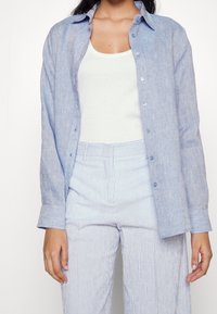 Femme portant une chemise bleu clair boutonnée sur un débardeur blanc et un pantalon à rayures bleu et blanc taille haute, debout devant un fond uni.
