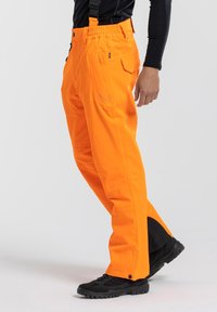 Heldere oranje sneeuwbroeken met verstelbare banden, meerdere zakken en een stoffen paneel bij de enkels voor extra bescherming. Zwarte schoenen zichtbaar.