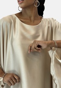 Beige satin topp med bred halsringning och avslappnad passform. Har delikat drapering och en mjuk, slät textur. Silverfärgade cirkulära örhängen visas.