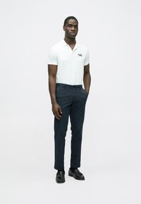 Polo branca com gola e logótipo, combinada com calças azul-marinho escuras. O modelo usa sapatos pretos e está em pé contra um fundo simples.