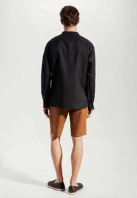 Hombre de pie de espaldas, vistiendo una camiseta negra de manga larga, pantalones cortos marrones hasta la rodilla y zapatos negros sin cordones sobre un fondo liso.