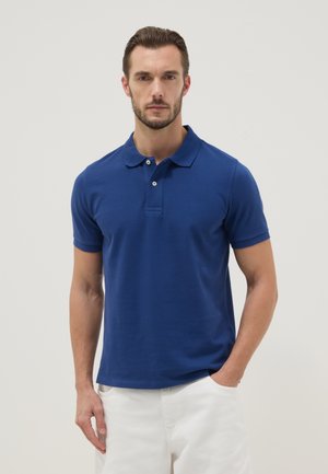 Mann trägt ein blaues kurzärmeliges Poloshirt und weiße Hose, steht mit einer Hand in der Tasche vor einem einfarbigen hellen Hintergrund.