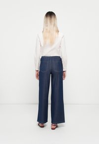 Pantaloni in denim a gamba larga con vestibilità rilassata, colore blu scuro, vita elastica e due tasche posteriori. Indossati con una camicia di colore chiaro.