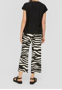 Femme portant un haut noir à manches courtes, un pantalon large à imprimé zèbre noir et blanc, et des sandales plateformes noires, se tenant devant un fond gris.