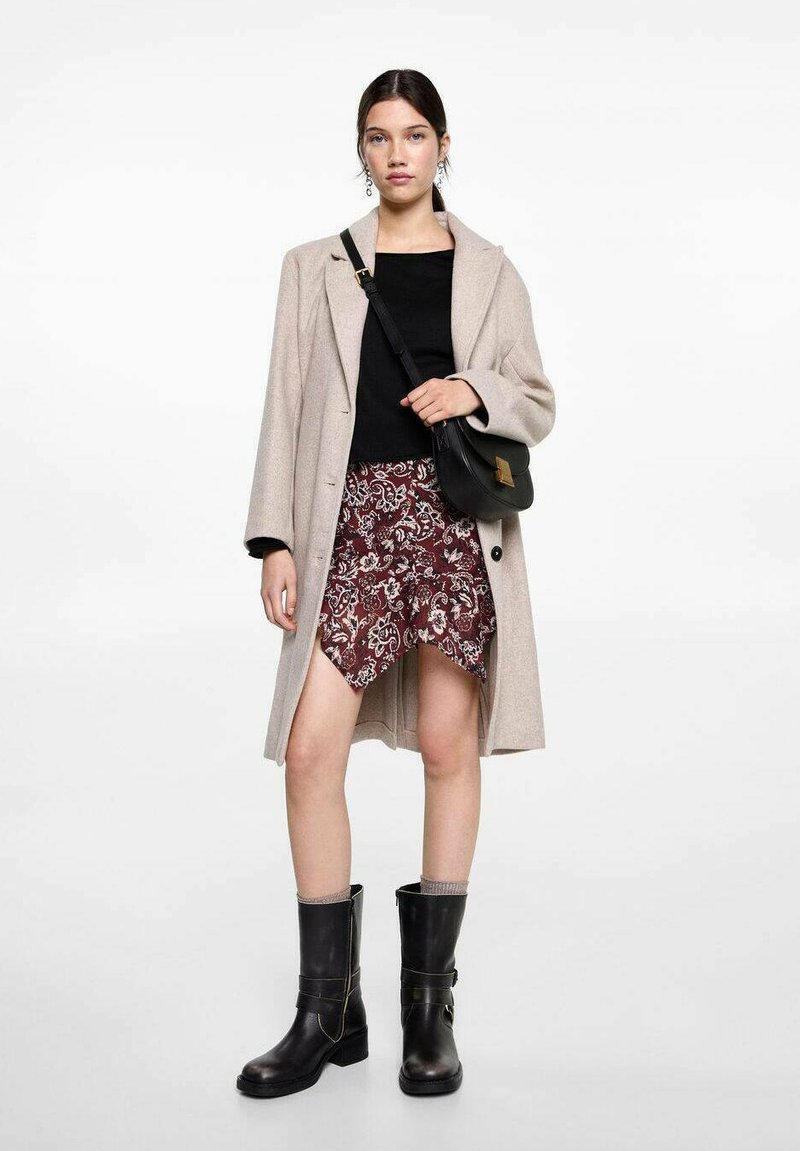 Beige Wollmischmantel, schwarzes Oberteil, bordeauxfarbenes gemustertes Minirock, schwarze kniehohe Stiefel mit Schnalle und Umhängetasche.