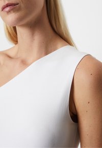 Haut blanc à une épaule fabriqué en tissu lisse. Il présente un design épuré avec un col incurvé et un côté légèrement ouvert, mettant en valeur la texture de la peau.