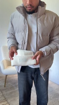 Személy által tartott fehér bordázott kötött sapka, hajtott mandzsettával és kis texturált logófelvarróval. A háttérben világos berendezési tárgyak láthatók.