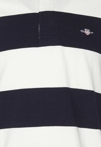 Polo a righe orizzontali bianche e blu navy con colletto parzialmente visibile e logo GANT ricamato sul petto.