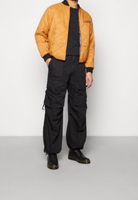 Veste bomber matelassée orange avec des accents noirs, associée à un pantalon cargo noir ample. Le mannequin porte des chaussures noires à lacets.