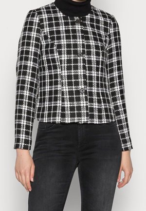 Black and white plaid button-up jacket worn over a black turtleneck, paired with high-waisted black jeans.