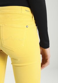 Primer plano de una persona que lleva puestos unos pantalones vaqueros amarillos brillantes con un bolsillo trasero y una camiseta de manga larga negra, de pie contra un fondo liso.