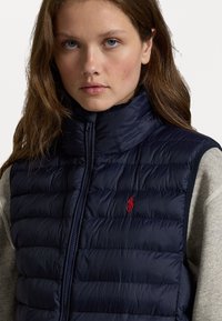 Vesta puffer de marin cu guler înalt, închidere cu fermoar și accent roșu al logo-ului. Material texturat, matlasat. Poartă peste o cămașă gri cu mâneci lungi.