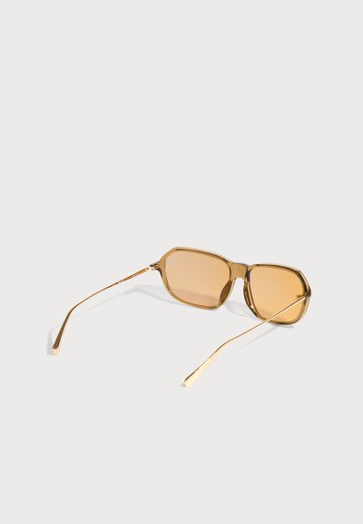 Lunettes de soleil rectangulaires transparentes marron clair avec des branches métalliques fines dorées sur un fond gris clair.