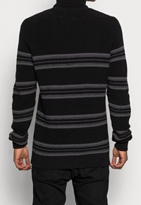 Maglione nero a maglia con strisce orizzontali grigie, con colletto alto e texture a coste. Maniche lunghe con polsini a coste.