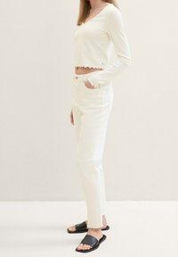 Valkoinen, ribbineulottu pitkähiha-paita, jossa on aaltoilevat reunat, yhdistetty valkoisiin suoralahkeisiin housuihin. Mustat liukusandaalit täydentävät asun.