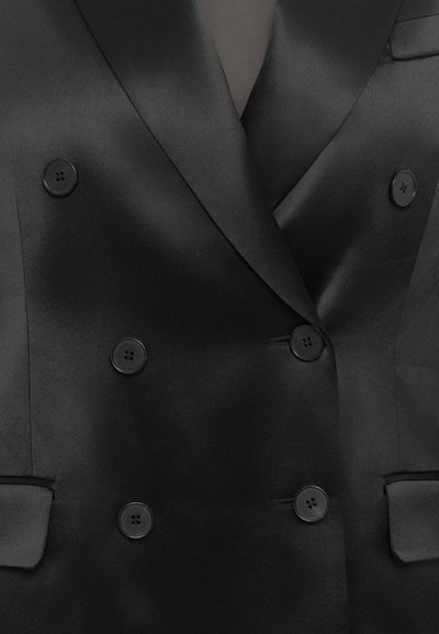 Zwarte satijnen double-breasted blazer met zes knopen en piekrevers over een zwart kledingstuk, met zicht op het bovenlichaam en details van de zakken.