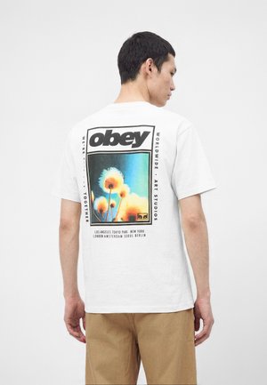 Hombre con una camiseta blanca con el logo "obey" y un gráfico de flor colorido en la espalda, combinado con pantalones beige, sobre un fondo sencillo.