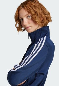 Marineblaue Reißverschlussjacke mit hohem Kragen, strukturiertem Stoff, mit weißen drei-Streifen-Akzenten auf den Schultern. Modell posiert mit verschränkten Armen.