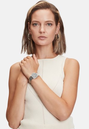 Montre-bracelet en argent avec un cadran rond, un fond noir et un bracelet en maille. Portée sur un poignet nu, complétée par un haut texturé de couleur claire.