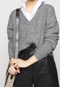 Maglione grigio in lana a V indossato su una camicia bianca con colletto, abbinato a pantaloni neri in pelle a vita alta. Include una borsa con texture.