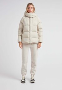 Puffjakke i lys beige med høj krave, store rektangulære quiltede sektioner og lange ærmer. Sammen med matchende træningsbukser og sneakers.