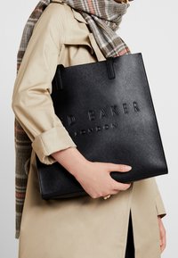 Czarna torba tote o fakturowanej powierzchni z wytłoczonym logo "Ted Baker London", z dwoma solidnymi uchwytami, trzymana na tle beżowego trencza.