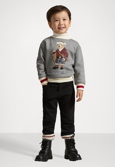 Sudadera gris con un gráfico de oso, cuello alto blanco, pantalones negros y botas negras con acentos en crema y rojo, de pie sobre un fondo sencillo.