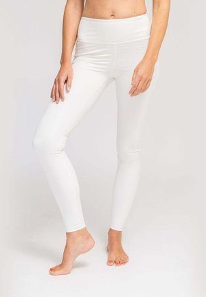 Frau trägt weiße Leggings mit hoher Taille, steht barfuß mit einem angewinkelten Bein und die Hände auf den Oberschenkeln auf einem einfarbigen Hintergrund.