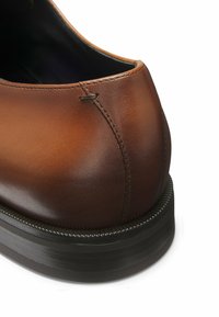 Chaussure en cuir marron avec une texture lisse, bout arrondi, détails cousus et semelle en caoutchouc noir. Aucun logo ou motif visible.