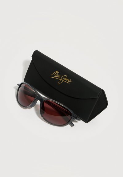 Μαύρα γυαλιά ηλίου Maui Jim με σκούρους κόκκινους φακούς τοποθετημένα δίπλα σε μαύρη σκληρή θήκη με χρυσό λογότυπο, σε λευκό φόντο.