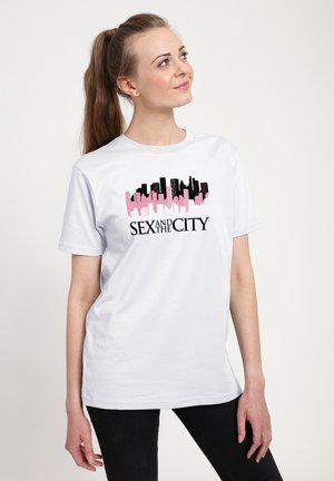 Ung kvinde med hestehale iført hvid T-shirt med rosa og sort bysilhuet og teksten "Sex and the City", kigger opad.